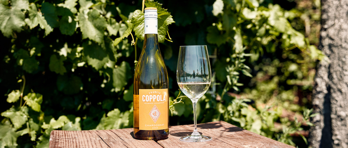 Weinflasche und Weinglas auf Holztisch vor grünem Blätterhintergrund.