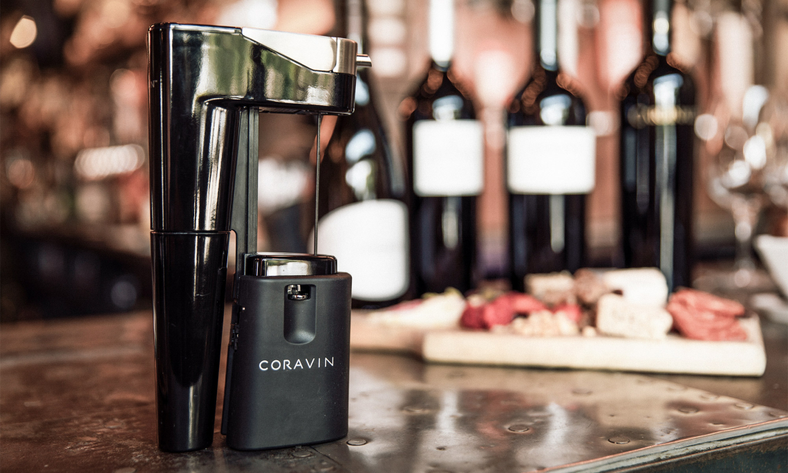 Weinausgießgerät vor unscharfem Hintergrund mit Weinflaschen und Käseplatte.