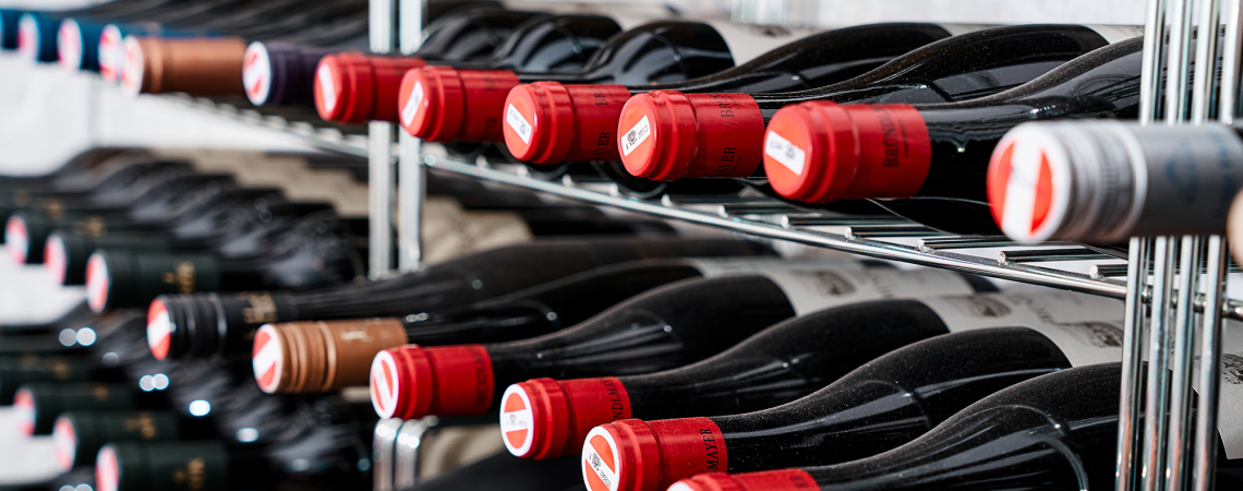 Nahaufnahme von horizontal gelagerten Weinflaschen mit roten Kapseln im Weinregal.