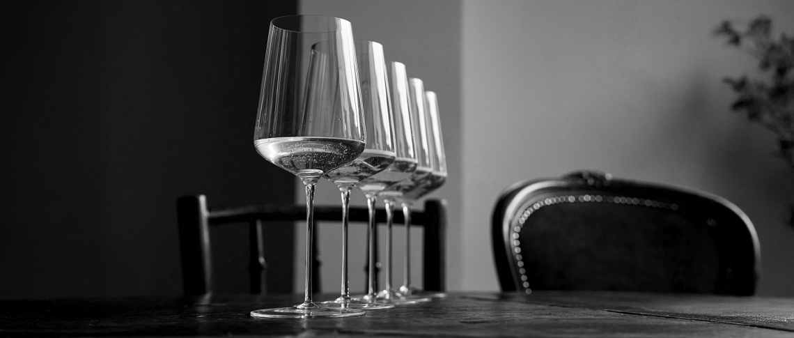 Vier elegante Weingläser auf Holztisch, im Hintergrund unscharfe Stühle.