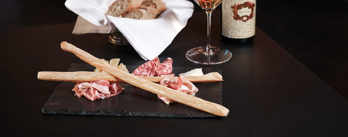 Wurst- und Käseplatte mit Brotstangen auf schwarzem Teller, begleitet von Wein.