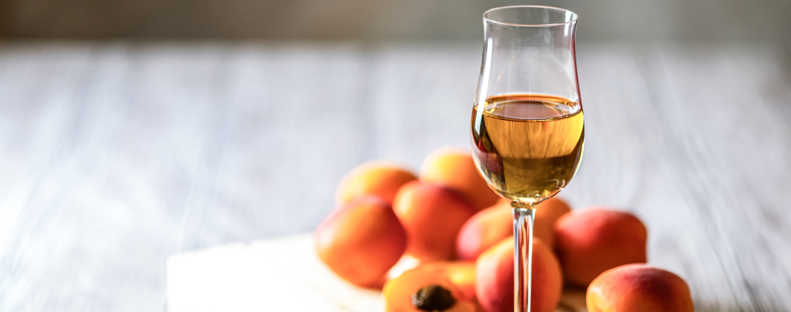 Ein Glas mit bernsteinfarbener Flüssigkeit, umgeben von frischen Aprikosen auf Holz.