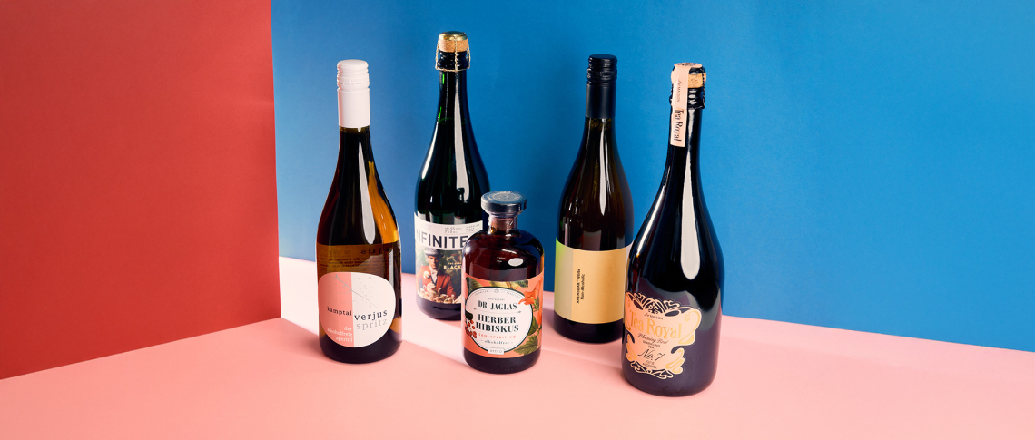 Fünf verschiedene Getränkeflaschen vor rot-blauem Hintergrund auf rosa Boden.