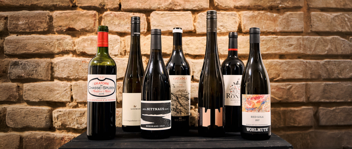 Sechs Weinflaschen vor einer rustikalen Ziegelwand auf einem dunklen Holztisch.