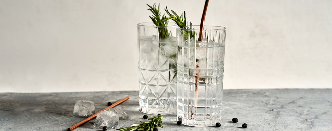 Zwei stilvoll gemusterte Gläser mit Gin Tonic und Rosmarin, Kupferstrohhalm, auf Marmortisch.