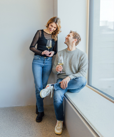Paar mit Weingläsern, lächelnd, neben einem großen Fenster; lässige Freizeitkleidung.