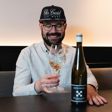 Mann mit Brille und Mütze hält ein Weinglas neben einer Weinflasche.