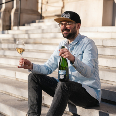 Mann sitzt auf Treppe, hält Weinflasche und -glas, trägt helle Jeansjacke, Basecap.