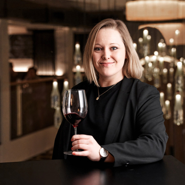 Frau in schwarzem Blazer hält Weinglas in elegantem, stimmungsvoll beleuchtetem Ambiente.