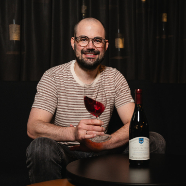 Mann mit Brille hält Weinglas neben Weinflasche, sitzt entspannt vor dunklem Hintergrund.