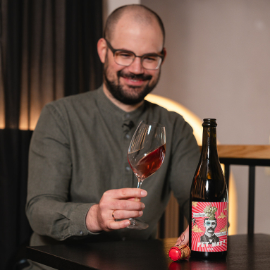 Mann mit Weinglas hält Flasche mit auffälligem Etikett auf Holztisch.