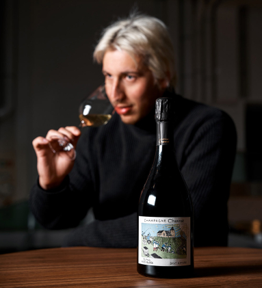 Champagnerflasche auf Holztisch, Person im Hintergrund riecht an Weinglas.