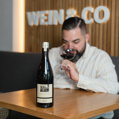 Weinflasche auf Tisch, Mann im Hintergrund genießt ein Glas Rotwein.