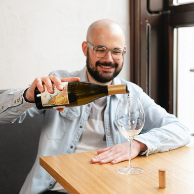 Mann schenkt Weißwein in ein Glas an einem Holztisch ein.