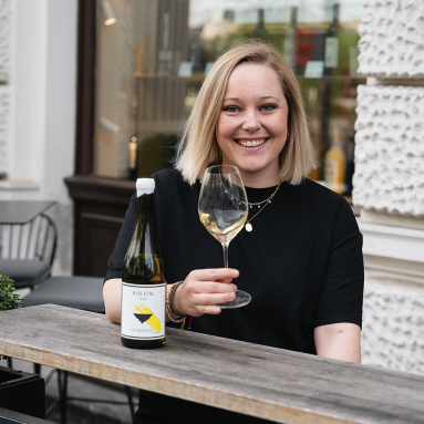 Frau lächelt mit Weinglas und Flasche auf rustikalem Tisch im Freien.