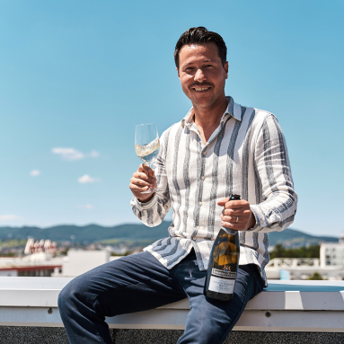 Mann in gestreiftem Hemd hält Weinglas und Flasche auf Dach mit Bergkulisse.