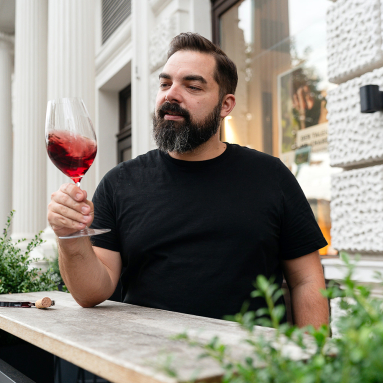 Mann in schwarzem T-Shirt hält Weinglas mit rotem Getränk draußen.