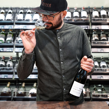 Mann in dunklem Hemd riecht an einem Weinglas, Weinflasche in der Hand.