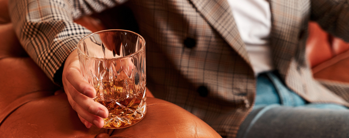 Person hält ein Kristallglas mit Whiskey auf einem braunen Ledersofa.
