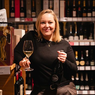 Frau in Weingeschäft, hält Weinglas und Flasche, lächelt in die Kamera.