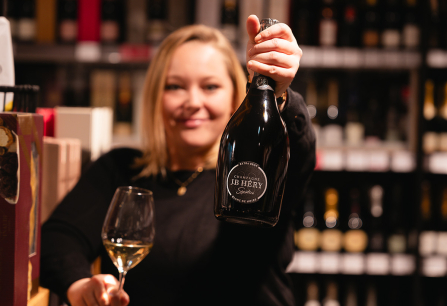 Frau präsentiert eine Champagnerflasche in einem Weinladen, lächelnd mit gefülltem Weinglas.