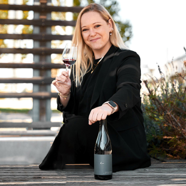 Frau in schwarzem Anzug hält Weinglas, Weinflasche auf Holzbank.