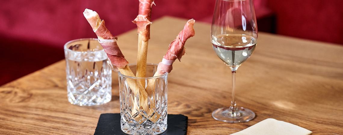 Knusprige Grissini mit Schinken in Kristallglas, begleitet von Wasser und Weißwein.