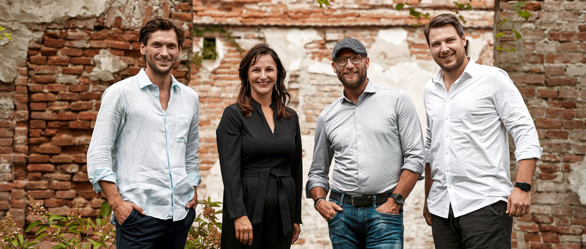 Vier Personen lächeln vor einer alten Backsteinmauer.