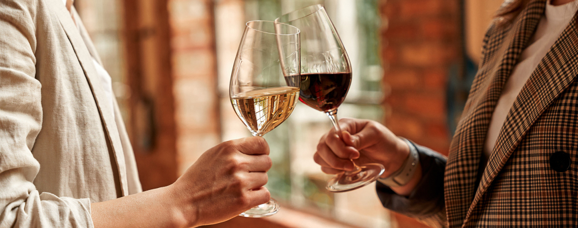 Zwei Personen stoßen mit Rot- und Weißweingläsern an, in stilvoller, warmer Umgebung.