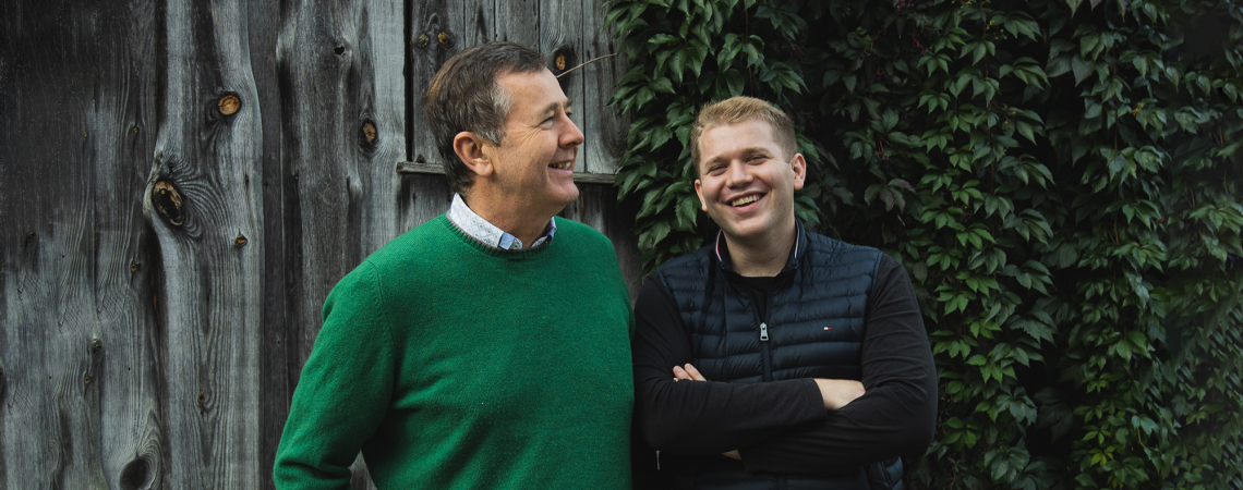 Zwei Männer lachend vor Holzwand und efeubewachsener Hauswand.