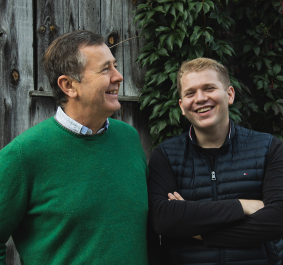 Zwei Männer lachend vor Holzwand und efeubewachsener Hauswand.