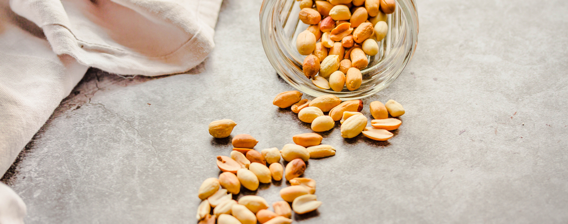 Umgekipptes Glas mit Erdnüssen auf grauem Untergrund und beige Stoff.