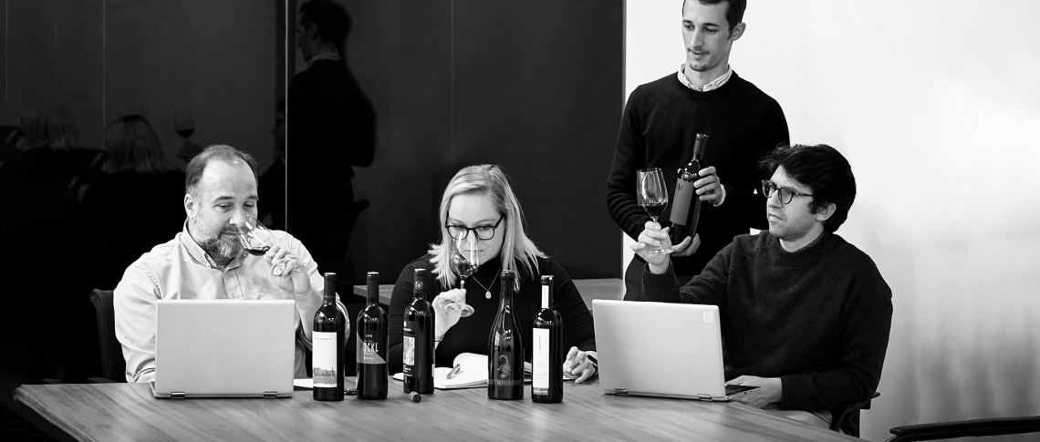 Vier Personen prüfen Weinflaschen und Weinproben an einem Tisch mit Laptops.