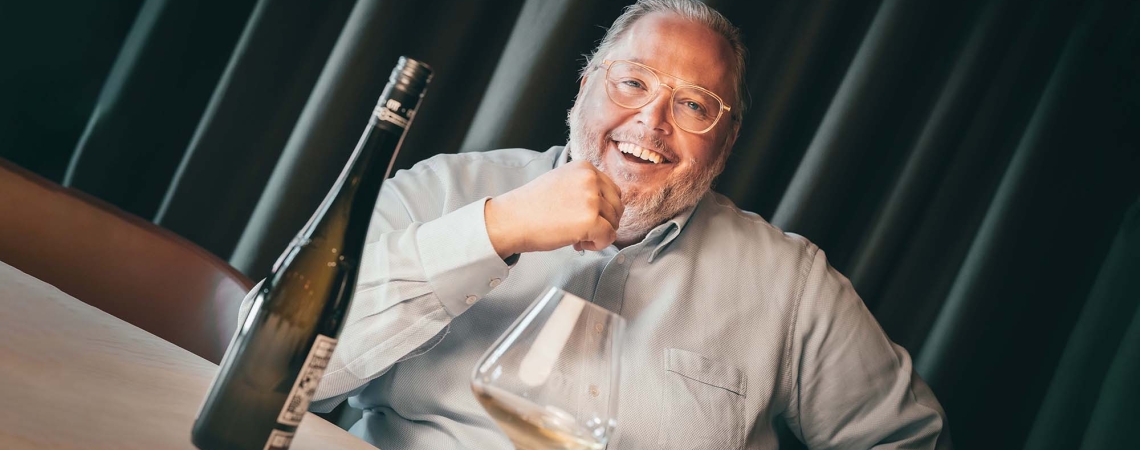 Mann sitzt lächelnd am Tisch mit Weinflasche und Glas davor.