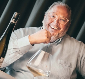 Mann sitzt lächelnd am Tisch mit Weinflasche und Glas davor.