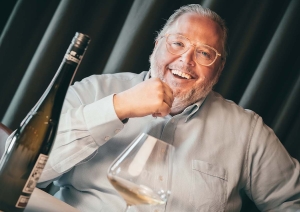 Mann sitzt lächelnd am Tisch mit Weinflasche und Glas davor.