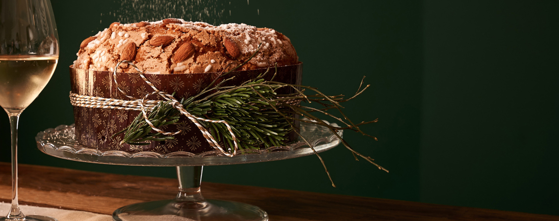 Festlicher Kuchen mit Mandeln und Tannenzweig-Dekoration auf Glasplatte neben Weinglas.