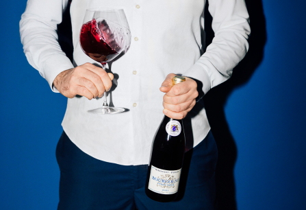 Person in weißem Hemd hält Rotweinglas und Weinflasche vor blauem Hintergrund.