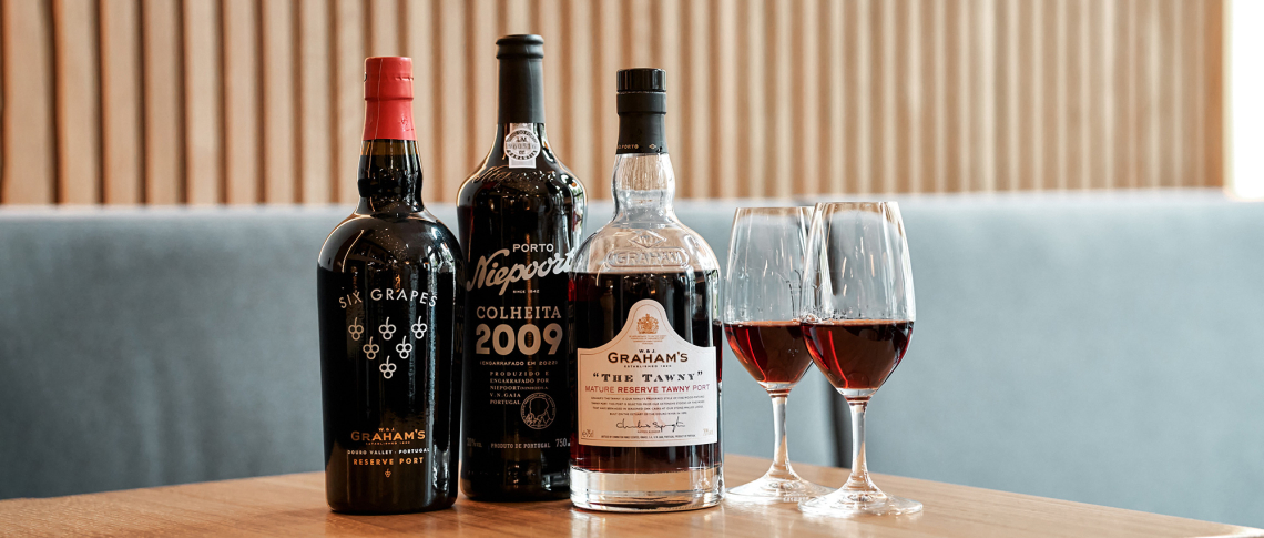 Drei Portweinflaschen und zwei Gläser auf Holztisch vor blauer Polsterbank.