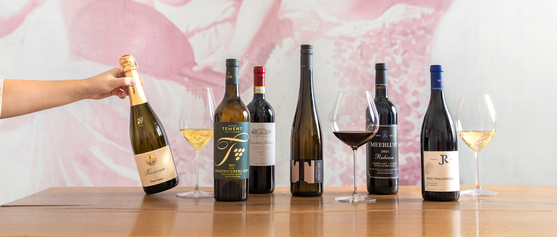 Fünf Weinflaschen und Gläser auf Holztisch vor rosafarbenem Hintergrund.