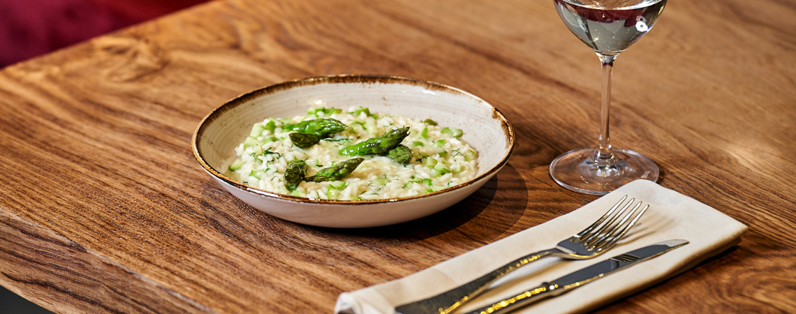 Risotto mit grünem Spargel auf rustikalem Holztisch, daneben Weinglas und Besteck.