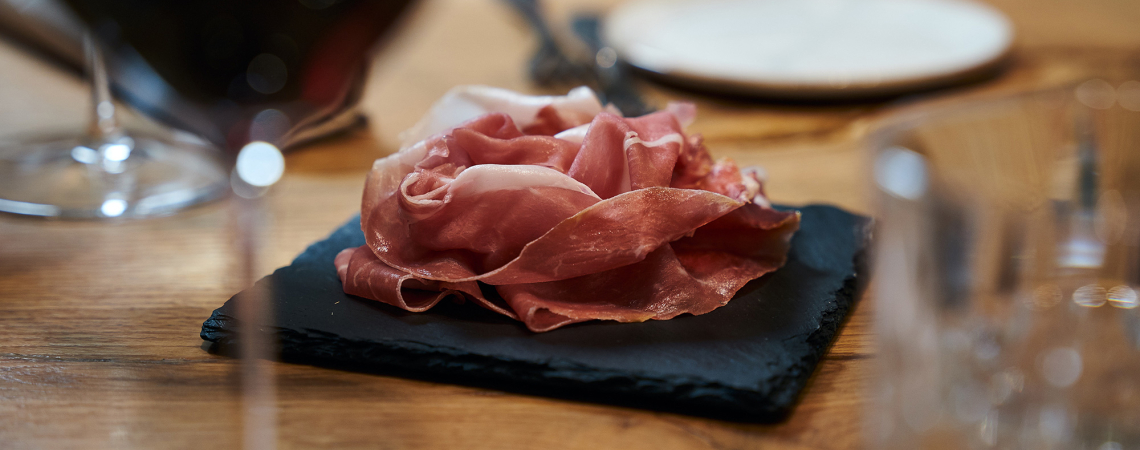 Dünne Schinkenstreifen auf einer schwarzen Schieferplatte, umgeben von Weingläsern.