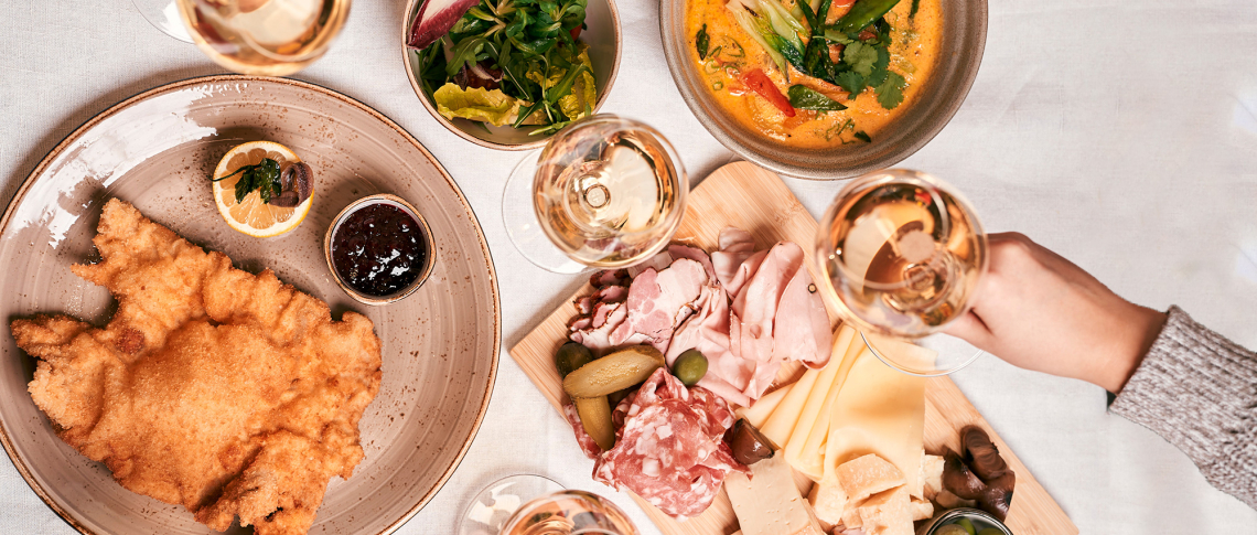 Tisch mit Wiener Schnitzel, Käse- und Wurstplatte, Salat, Curry und Weißweingläsern.