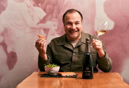 Lachender Mann mit Weinglas, Speisen und Flasche an Holztisch vor rosa Wand.