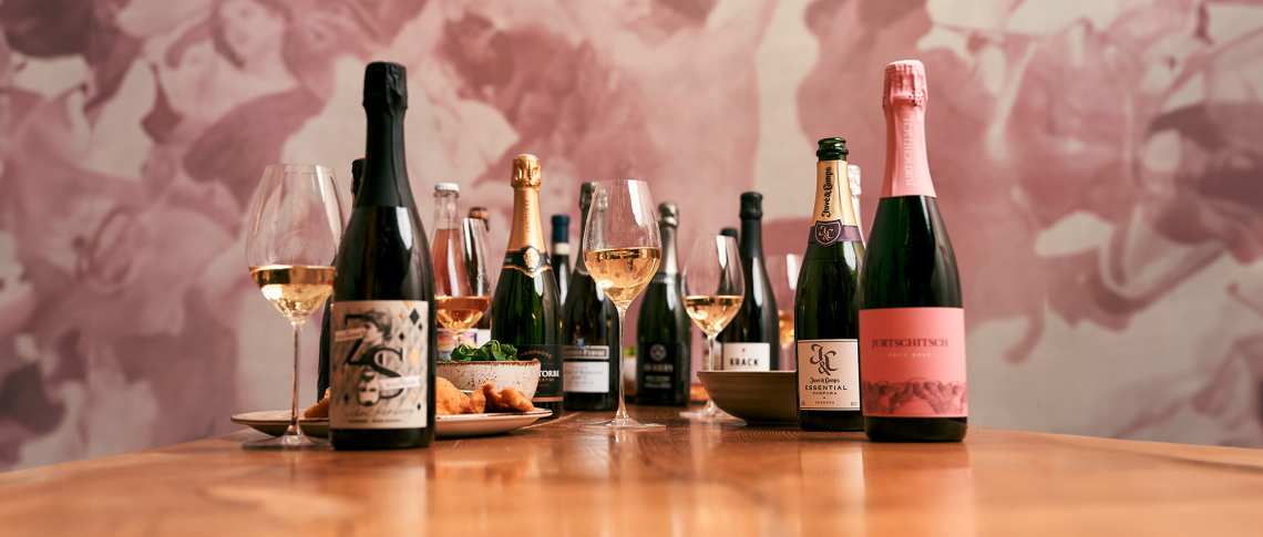 Verschiedene Schaumweinflaschen und Gläser auf Holztisch vor rosafarbener Kunstwand.