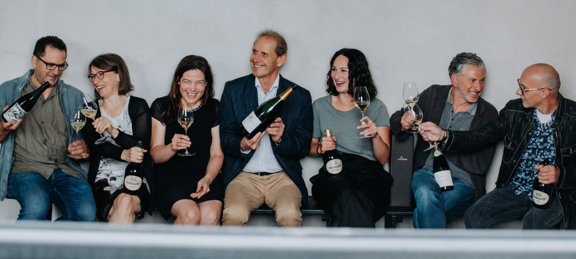 Sieben lachende Personen mit Weinflaschen und Gläsern sitzen auf einer Bank.