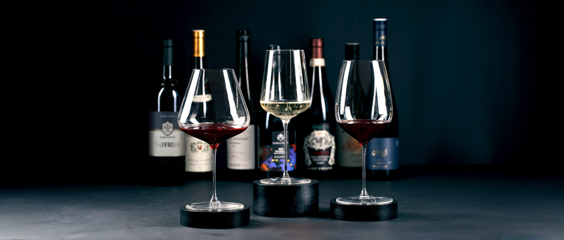 Drei Weingläser auf Podesten, im Hintergrund mehrere Weinflaschen vor dunklem Hintergrund.
