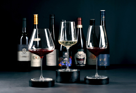 Drei Weingläser vor verschwommenen Weinflaschen auf dunklem Hintergrund.