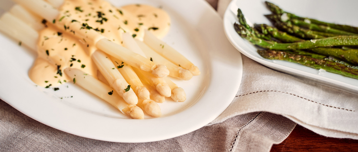 Weiße Spargel mit Sauce, garniert mit Kräutern, neben grünem Spargel auf Teller.