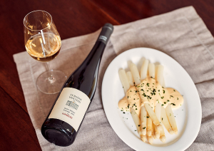 Weißwein neben Spargel mit Sauce auf weißem Teller, Holzuntergrund.